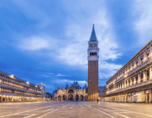 Vue panoramique de la place Saint-Marc à Venise avec la basilique Saint-Marc, le campanile et des pigeons au premier plan sous un ciel bleu.