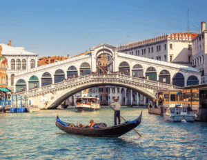Vue du Pont du Rialto en pierre enjambant le Grand Canal à Venise, avec des gondoles et des vaporettos sur l'eau.