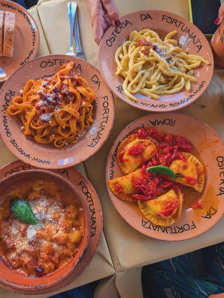 Photographie culinaire montrant une table de restaurant avec quatre spécialités italiennes différentes. Les assiettes portent l'inscription des villes "Roma, Milano, Bologna, Miami". L'image met en avant des produits frais, du basilic et des textures gourmandes.